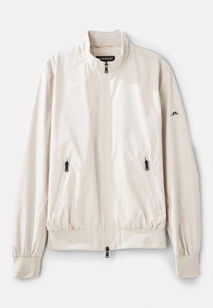Vaalean beige bomber-takki, valmistettu sileästä, vettä hylkivästä materiaalista. Ominaisuuksiin kuuluu pystykaulus, kaksi vetoketjullista taskua ja elastiset hihansuut.