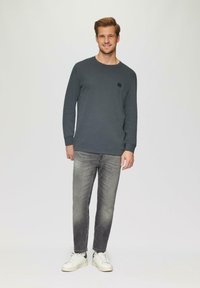 Graues Langarmhemd mit kleinem quadratischem Logo auf der Brust, kombiniert mit ausgewaschenen grauen Jeans und weißen Turnschuhen. Schlichter Hintergrund.