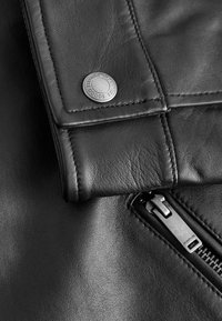 Detail einer schwarzen Lederjacke mit einem Metall-Druckknopf und Reißverschluss, glatte Textur, Nahaufnahme von Nähten und Nahtlinien.