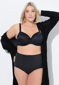 Zwarte satijnen bh met gemoldde cups en verstelbare straps, gecombineerd met hooggetailleerde zwarte slipjes met een klein strikdetail. Gebreide cardigan is over de schouders gedrapeerd.