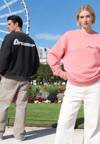 Homme en sweat-shirt noir avec "Dreamer" dans le dos regardant vers la gauche, femme en sweat-shirt rose "Mira Paris" regardant devant dans un parc avec une grande roue derrière.