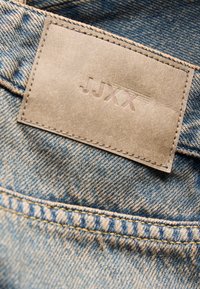Beiger Lederpatch mit eingeprägtem "JJXX" Markenlogo, auf blauen Denimstoff einer Jeans genäht.