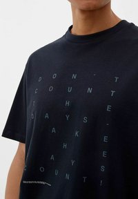 Schwarzes Baumwoll-T-Shirt mit einem geometrischen Textdesign in Dunkelgrün, das auf der Vorderseite "ZÄHLE DIE TAGE NICHT, MACH DIE TAGE ZÄHLEN!" zeigt.