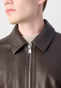 Brun læderjakke med en tekstureret overflade, en klassisk krave og en sølvfarvet lynlås. Ribbede sweater under tilføjer kontrast.
