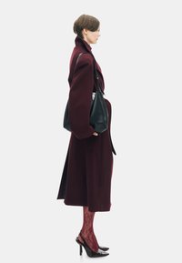 Bordeaux wollen jas met oversized mouwen, gecombineerd met een zwarte handtas. Rode gestreepte panty’s en zwarte gepolijste hakken maken de outfit compleet.