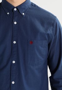 Mörkblå knapp-up skjorta i texturerad tissu med en ficka som har en liten röd broderad logotyp och vita knappdetaljer.