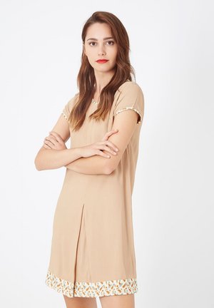 Mujer joven con cabello largo castaño, vestida con un vestido beige de mangas cortas y adornos estampados, de pie con los brazos cruzados frente a un fondo blanco liso.