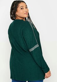 Maglione a maniche lunghe verde scuro con una vestibilità relaxed, caratterizzato da dettagli argentati lucidi lungo le maniche e una texture morbida.