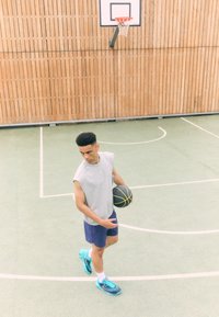 Grå ermeløs skjorte, marineblå shorts og lyseblå joggesko; holder en svart basketball på en grønn bane med trevegg i bakgrunnen.