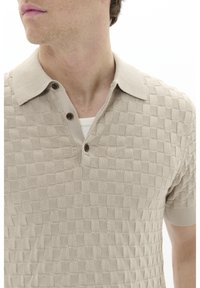 Beige adīts polo krekls ar strukturētu košer rakstu, īsām piedurknēm, apkakli un trim tumšām pogām pie kakla.