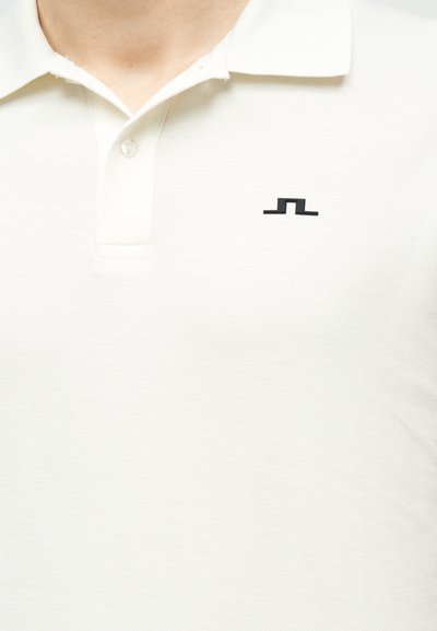 Άσπρο πόλο πουκάμισο με λευκό γιακά, δύο κουμπιά και ένα μικρό μαύρο γεωμετρικό λογότυπο στο αριστερό μέρος του στήθους.