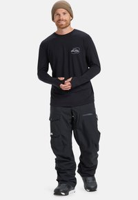 Zwarte lange mouwen shirt met een grijs logo en zwarte sneeuwbroeken met cargozakken. De persoon draagt een beige gebreide muts en donkere laarzen.