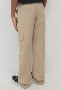 Beige Cargo-Hosen mit lockerer Passform, ausgestattet mit zwei Gesäßtaschen, einer Seitentasche und dezenten Nähedetails. Tragen mit schwarzen Schuhen.