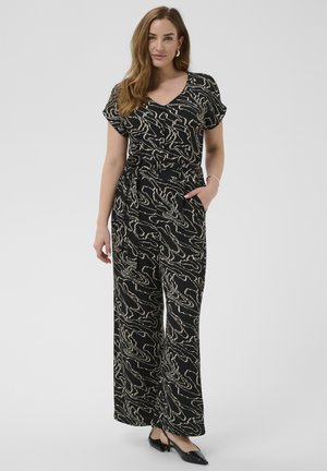 Kvinde stående med hånd i lommen, iført sort og beige kortærmet top med abstrakt mønster og bukser med vide ben samt sorte slingback-sko.