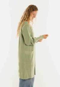 Vrouw met lang krullend haar, gekleed in een lichtgroene gebreide cardigan en blauwe jeans, terwijl ze de mouw van haar rechterarm aanpast.