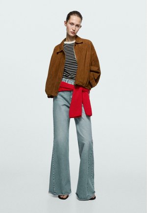 Frau trägt eine braune Wildlederjacke über einem schwarz-weiß gestreiften Hemd, hellblaue Jeans mit weitem Bein und einen roten Pullover, der um ihre Taille gebunden ist.