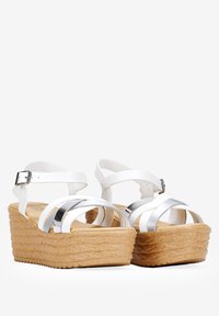 Sandales blanches à talon compensé avec une plateforme beige texturée, des accents argentés et des sangles croisées ajustables avec une fermeture à boucle.