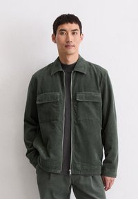 Zielona kurtka sztruksowa z przednim suwakiem, dwoma kieszeniami na piersi oraz kołnierzem. Noszona na czarnej koszulce, z teksturowanej tkaniny i o luźnym kroju.