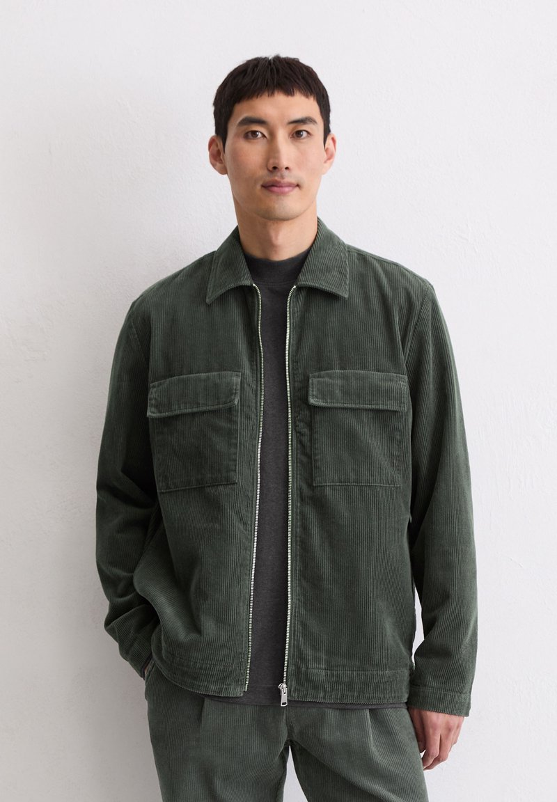 Zielona kurtka sztruksowa z przednim suwakiem, dwoma kieszeniami na piersi oraz kołnierzem. Noszona na czarnej koszulce, z teksturowanej tkaniny i o luźnym kroju.