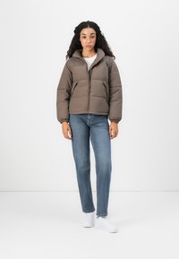 Donna con capelli ricci che indossa un giubbotto imbottito marrone, jeans blu e sneakers bianche, in piedi su uno sfondo bianco.