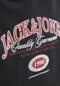 Tissu noir avec un texte audacieux en blanc et bordeaux. Comprend "JACK & JONES", "Vêtements de qualité" et un logo circulaire "1990".