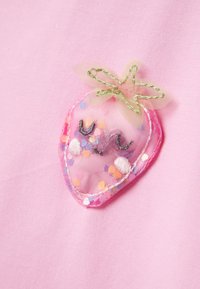 Roze stof heeft een doorschijnende aardbeitje applicatie met meerkleurige stippen, een groene bladaccent, en omrandde stiksels als detail.