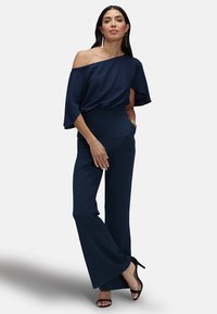 Navyblauw off-shoulder top met vloeiende mouwen, gecombineerd met op maat gemaakte flared trousers. De stof is glad, met een slanke silhouette en minimale details.