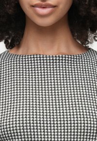 Schwarz-weißer Hahnentrittmuster-Stoff mit glatter Textur und einem Bootsausschnitt, umrahmt von lockigem Haar und Hauttönen.