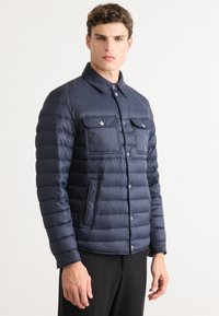 Moški oblečen v temno moder prešiti plašč z zaklopnimi gumbi in žepoma na prsih, stoji ob belem ozadju.