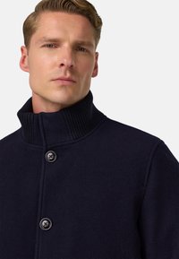 Námořnický vlněný kabát s ribbovaným límcem, obsahující dva velké knoflíky a strukturovaný design. Hladká textura a čisté linie.