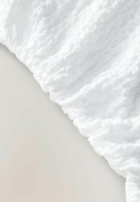 Bílá texturovaná tkanina se zvrásněným vzorem, vyrobená z měkkého, lehkého materiálu, volně visící, vytvářející jemné vlny.