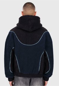 Marinblå fleecehuva med en texturerad yta, featuring ljusblå detaljerade sömmar och en stor huva. Baksidan visar ett böjt mönster.