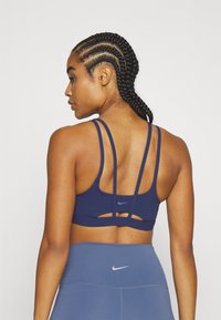 Mornarica sportski grudnjak s višestrukim tankim trakama, prorezima i Nike logotipom. Model ima pletenice i nosi usklađene plave hlače visoko strukirane.