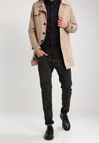 Man i beige trenchcoat, svart skjorta, mörka cargo byxor och svarta dressade skor, poserande mot en enfärgad ljus bakgrund.