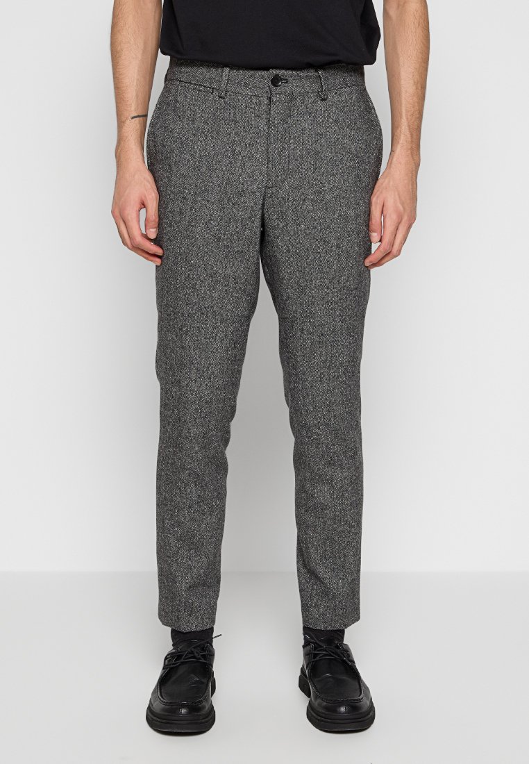 Pantaloni grigi a spina di pesce con vestibilità su misura. Presentano una chiusura con bottone e tasche laterali. Indossati con scarpe nere su uno sfondo neutro.