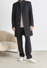 Lange zwarte jas met hoge kraag, textuurstof, voor knopen, gedragen over een beige shirt en donkere broek, gecombineerd met witte sneakers.
