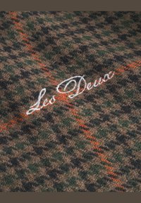 Tela marrón y verde con un diseño de pata de gallo; líneas naranjas y detalle bordado en blanco del logo "Les Deux". Textura suave.