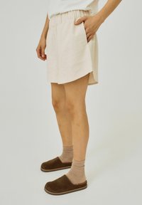 Light beige shorts with an elastic waistband, side pockets, and a slightly curved hem. Paired with brown slip-on shoes and light socks.