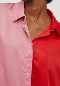 Camicia a blocchi di colore con una texture liscia e lucida, con un lato rosa e un lato rosso; design con colletto e tonalità a contrasto.