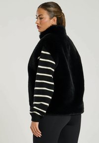 Svart fuskpälsväst med hög krage, i kombination med en långärmad randig svart och vit skjorta. Mjuk textur, avslappnad stil, sidovy.