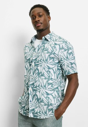 Mann trägt ein grün-weißes, kurzärmeliges Hemd mit Blattmuster und weißem Unterhemd, steht mit den Händen in den Taschen vor weißem Hintergrund.