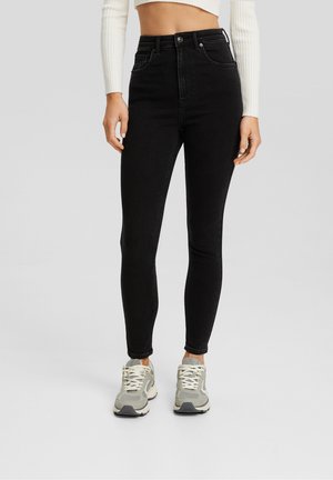 Femme portant un jean skinny noir taille haute, un pull court côtelé blanc, et des baskets grises et blanches sur un fond clair uni.