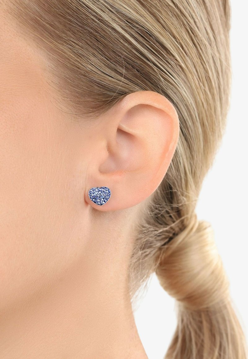 Hjärtförsedda stiftörhängen med blå glittrande yta, inlagda i en metallisk bas, bärs på örsnibben mot en bakgrund av rakt hår.