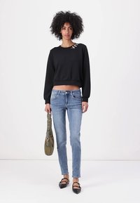 Melna īsā kapuce ar balonu piedurknēm, zili šaurie džinsi, melnas plakanās kurpes ar loku akcentiem un zaļa plecu soma.