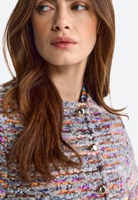 Texturierte, mehrfarbige Tweedjacke mit metallischen Knöpfen, die lila, orange und graue Fäden sowie ein gewebtes Detail am Kragen aufweist.