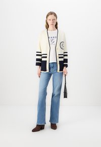 Cardigan crem cu dungi bleumarin, cu butoni aurii și patch cu logo. Împerecheat cu un tricou alb și blugi evazați de culoare albastru deschis. Botine maro cu toc.