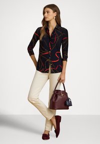 Sieviete stāv, ģērbusies melnā krekliņā ar sarkaniem un zelta ķēžu rakstiem, valkā bēšas bikses, bordo slofkas un nēsā bordo rokassomu ar pievienotu nelielu melnu somiņu.