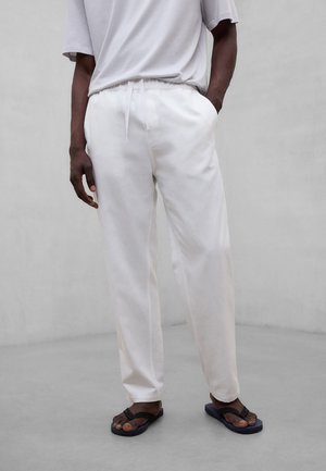 Person iført løstsittende hvite bukser med snor og svarte flip-flops, stående med én hånd i lommen mot en ensfarget bakgrunn.