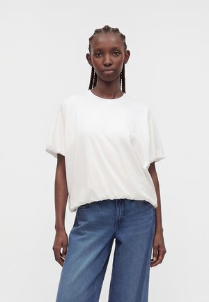Ung kvinde med fletninger, iført en løs hvid kortærmet top og blå højttaljede jeans, stående mod en ensfarvet baggrund.