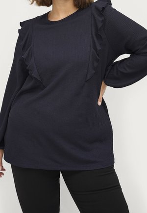 Mujer con una prenda superior oscura de manga larga con detalles de volantes en los hombros y pantalones negros ajustados, con una mano en la cadera.